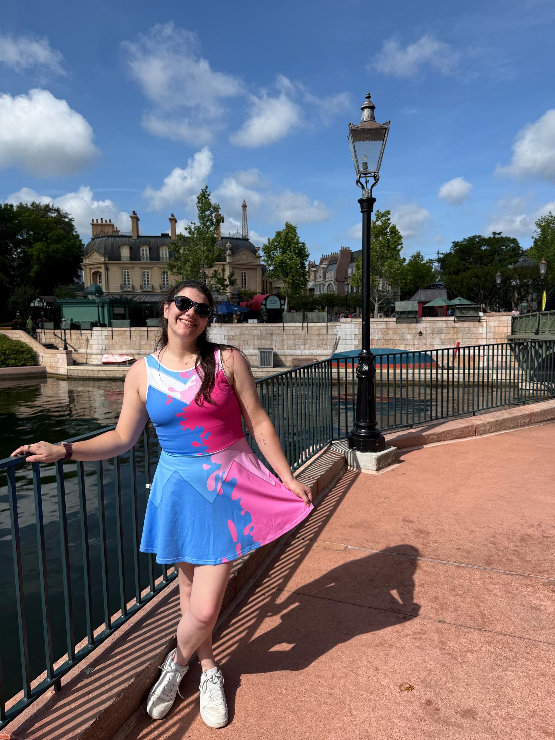 May include: A sleeveless crop top and skater skirt set in blue and pink with a paint-splatter design. The outfit is worn with white sneakers. The background includes a body of water, buildings, and a blue sky with clouds.